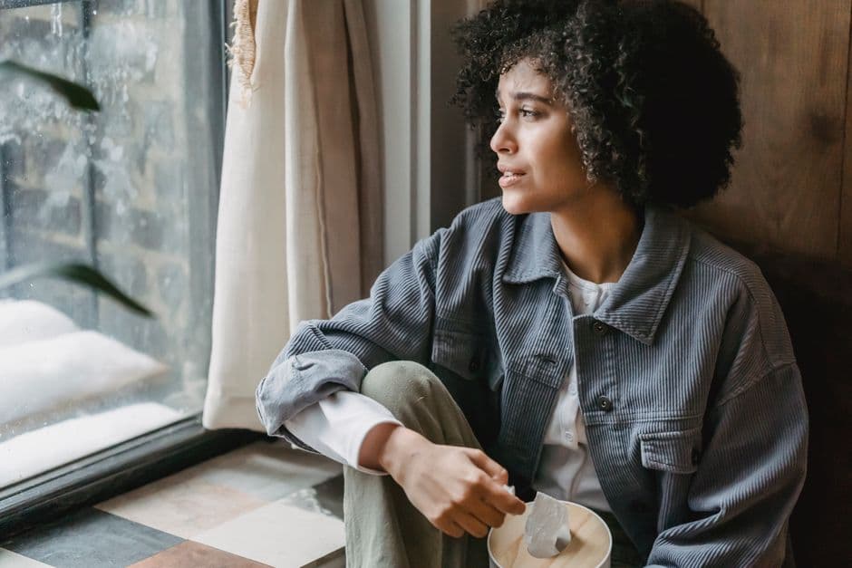 High angle of upset African American female suffering from loss and misfortune sitting on windowsill and looking away